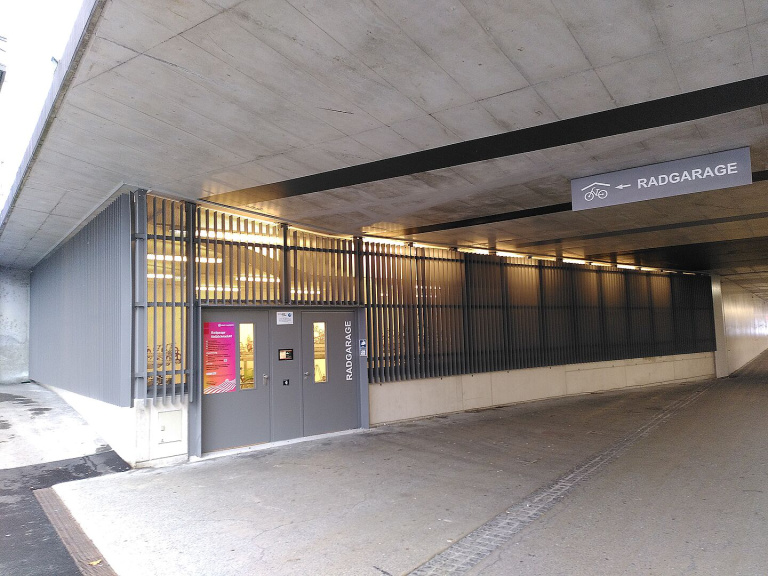 Eingang der Radgarage Nelböckviadukt unter einem Betonviadukt mit Beschilderung und beleuchteten Fahrradständern