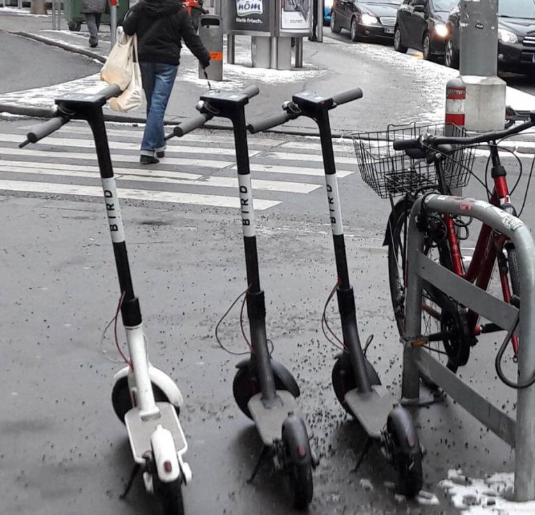 Drei E-Scooter stehen an einer Fußgängerzone in Salzburg, Radfahrer im Hintergrund.
