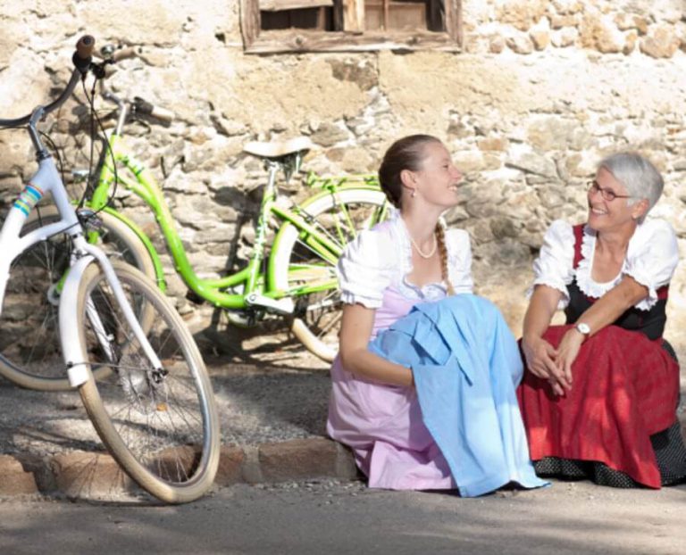 Zwei Frauen in Trachten sitzen lächelnd neben Fahrrädern vor einer Steinmauer.