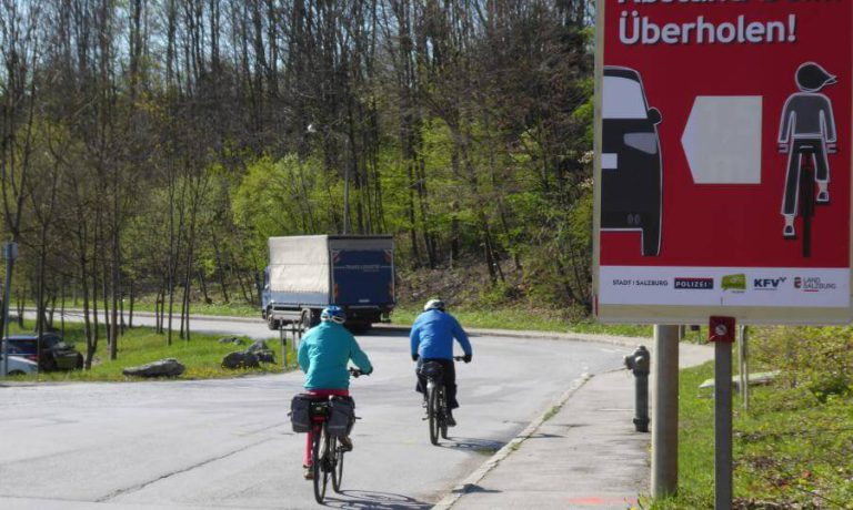 Zwei Radfahrer fahren an einem Verkehrsschild vorbei, das zum Überholen auffordert.