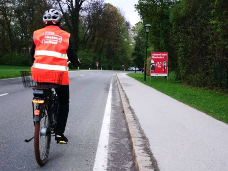 Radfahrender mit Warnweste auf Straße, Info-Plakat zu Sicherheitsabstand von 1,5 m.