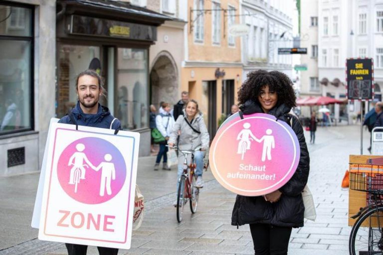 Zwei Personen halten Schilder für die Aktion „Schaut aufeinander“ in einer Fußgängerzone.