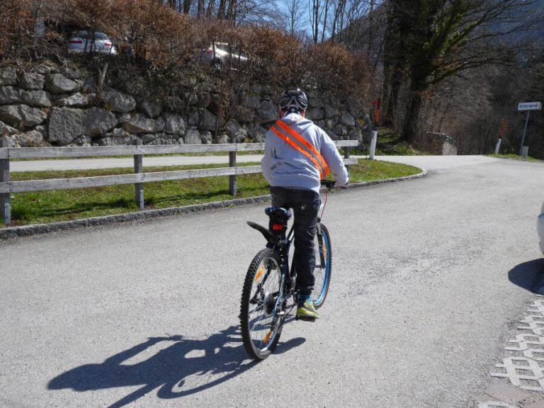 Kind auf dem Fahrrad mit Reflexschärpe fährt eine Straße entlang im Freien.