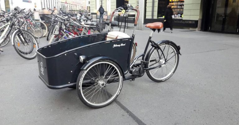 Schwarzes E-Lastenrad mit braunen Details auf einer Stadtstraße, umgeben von anderen Fahrrädern.