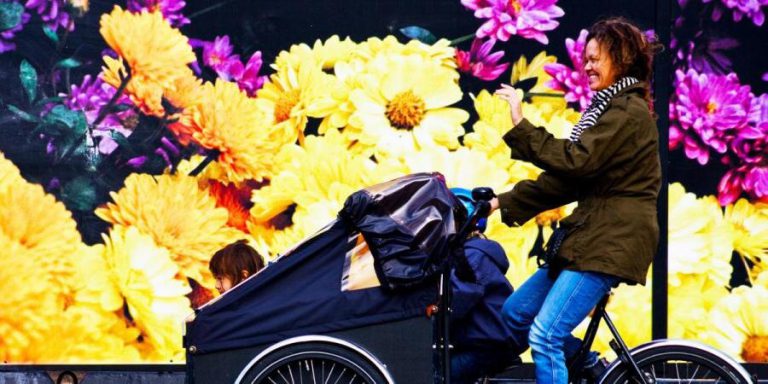 Familie fährt fröhlich mit einem Lastenrad vor buntem Blumenhintergrund.
