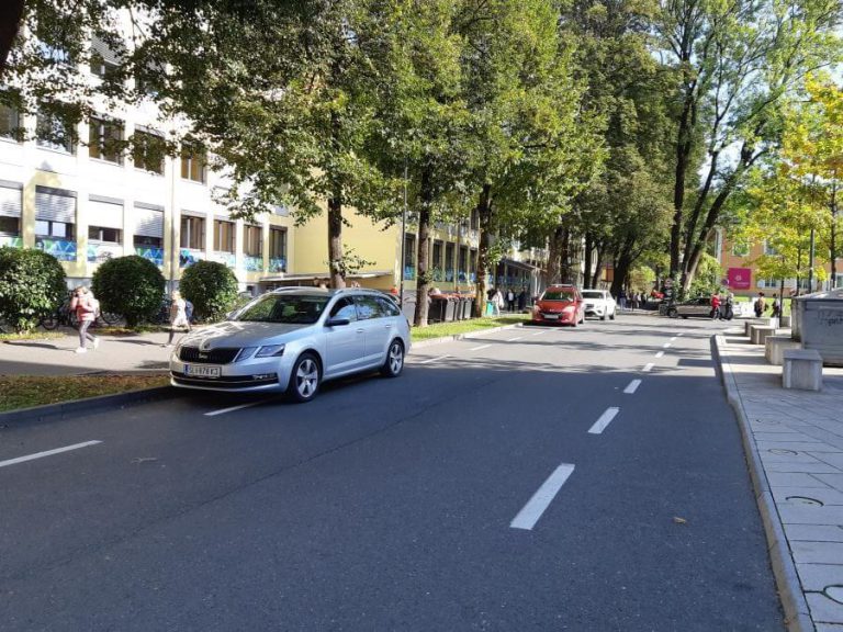 Straße mit Fahrzeugen und Passanten, umgeben von Bäumen und Gebäuden.