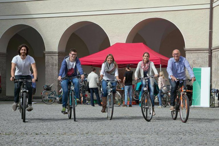 Fünf Radfahrer fahren lächelnd auf einem Platz vor einem Zelt.