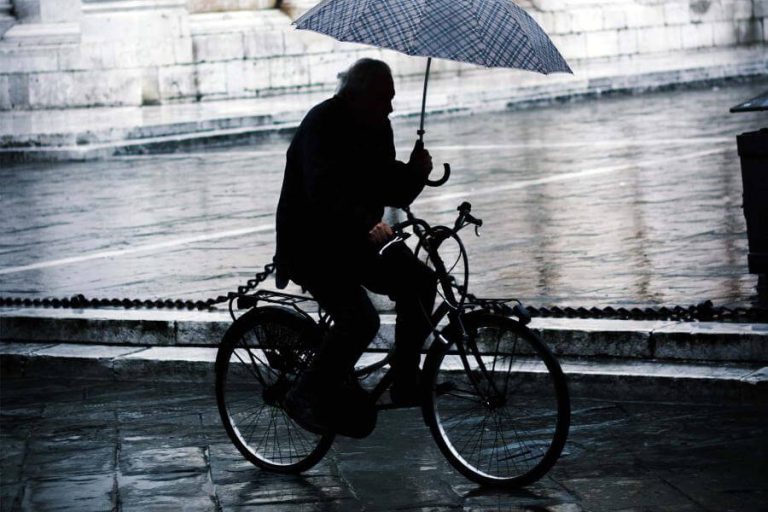 Radfahrer mit Regenschirm fährt bei Nieselregen durch eine Stadtstraße.