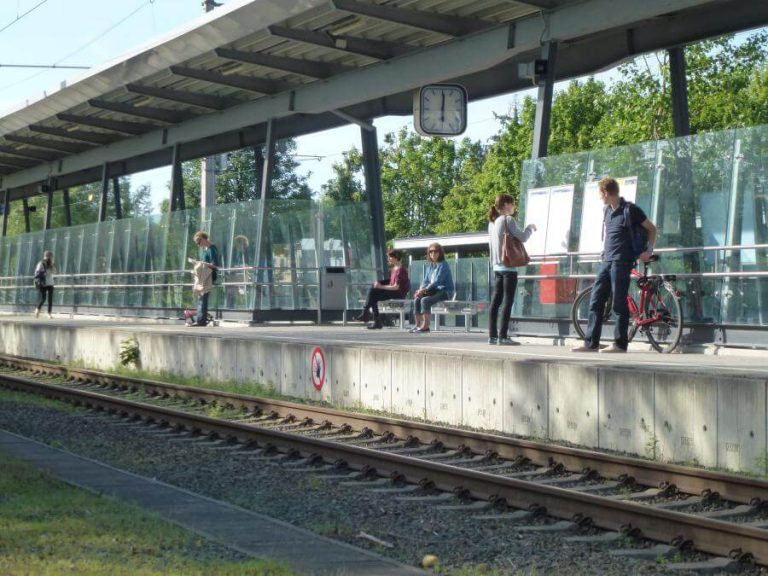 Fahrgäste warten am Bahnhof, einige mit Fahrrädern, in sonniger Umgebung in Salzburg.