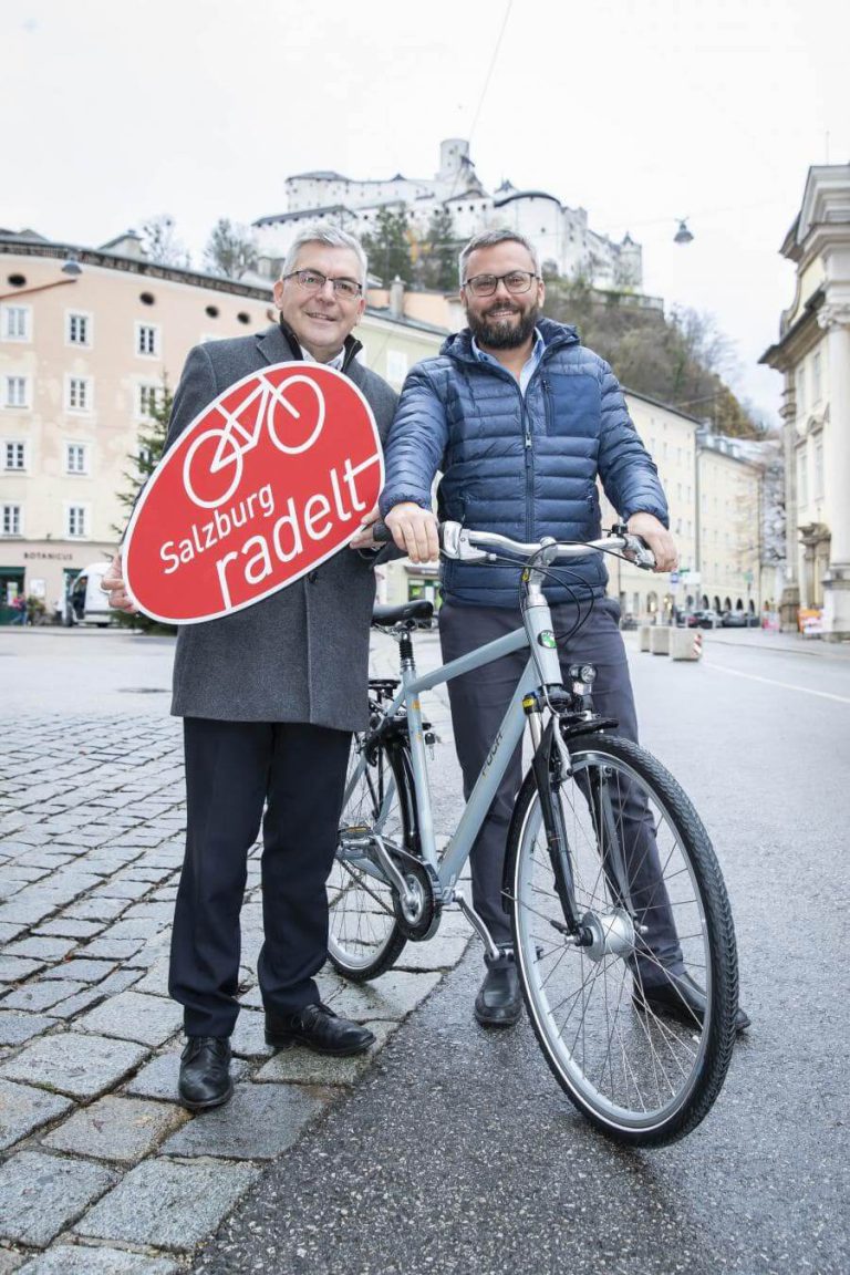 Zwei Männer halten ein Schild „Salzburg radelt“ und stehen neben einem Fahrrad.