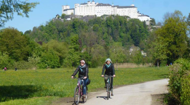 Zwei Frauen radeln entspannt auf einem Weg mit Blick auf Burg Hohensalzburg.