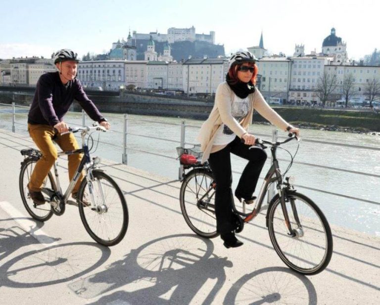 Frau und Mann radeln entlang der Salzach, im Hintergrund die Altstadt von Salzburg.