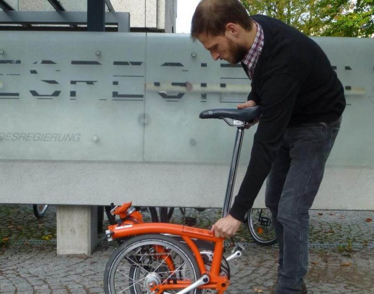 Mann faltet ein orangefarbenes Fahrrad vor einer Wand in Salzburg.