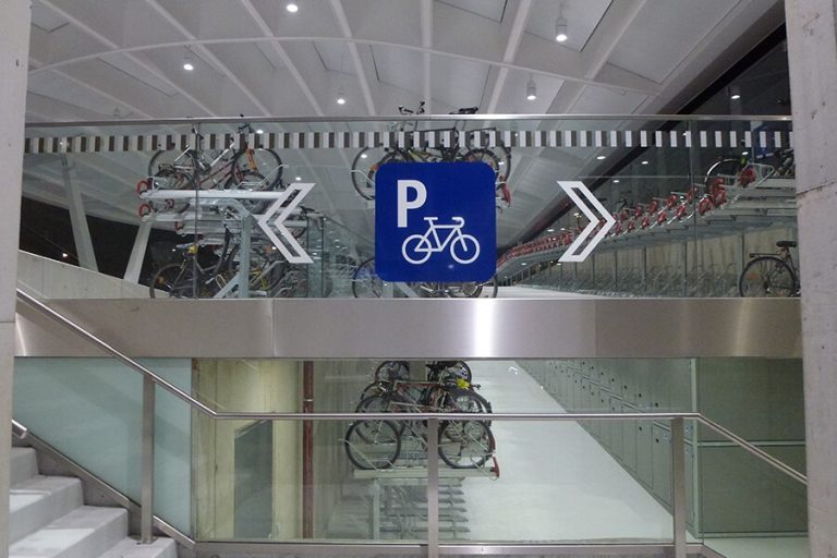 Fahrradparkplatz mit Schildern, moderne Architektur und stapelbare Fahrradständer.