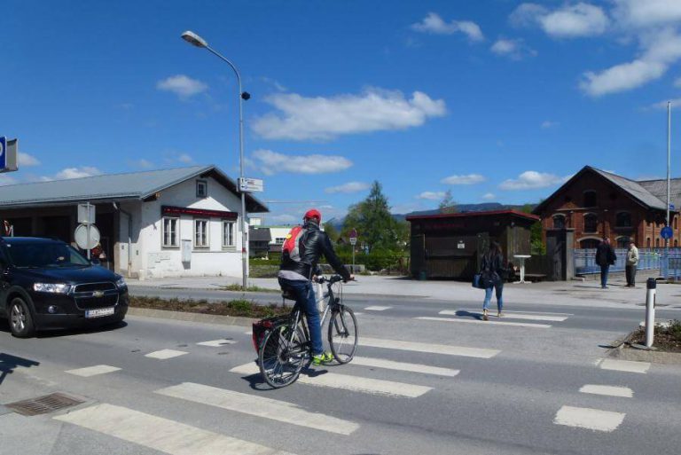 Fahrradfahrer kreuzt eine Straße mit Fußgängern, Umgebung zeigt eine ländliche Szenerie.