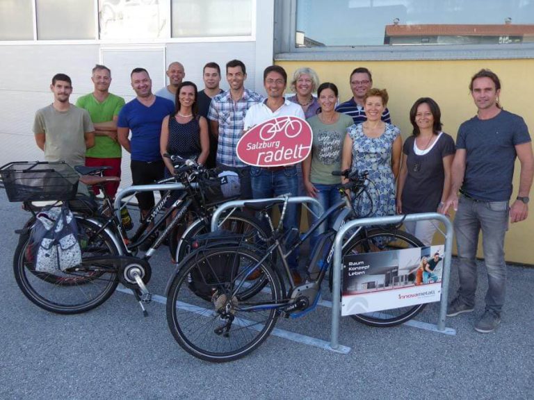 Gruppe von Radfahrern mit Fahrrädern und dem Schild „Salzburg radelt“ vor Gebäude.