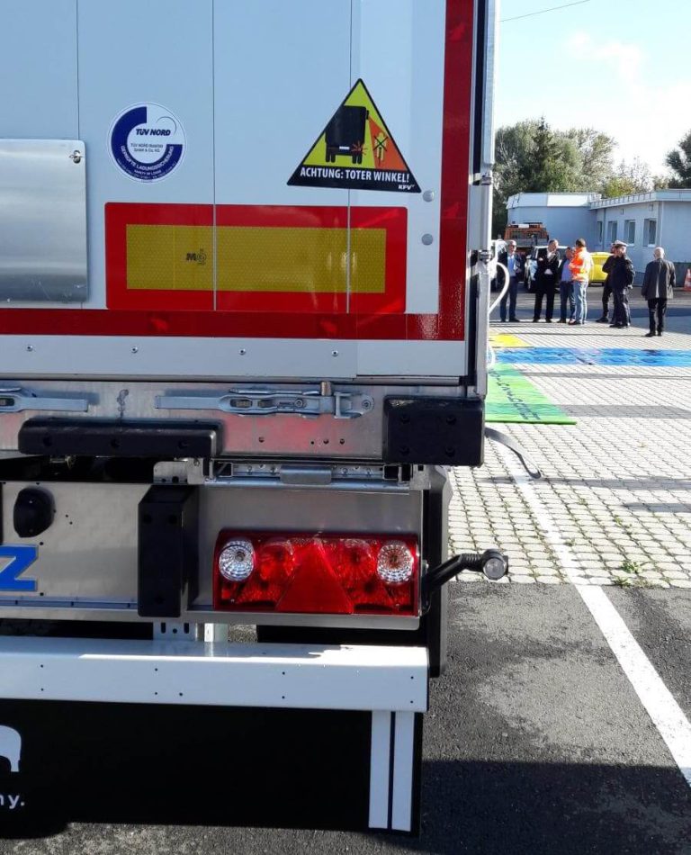 Warnhinweis „Achtung: Toter Winkel“ auf einem LKW, im Hintergrund Personen bei einer Veranstaltung.