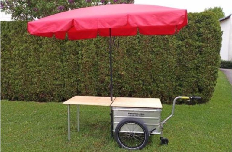 Fahrradanhänger mit rotem Sonnenschirm und Tisch für Veranstaltungen in Salzburg.
