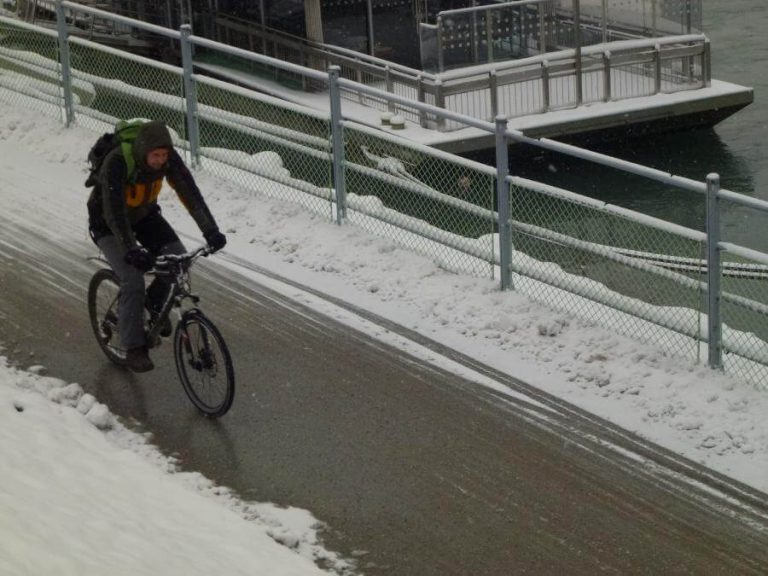 Radfahrer mit Rucksack fährt auf snowy Weg entlang einem Gewässer. Winterliche Bedingungen.