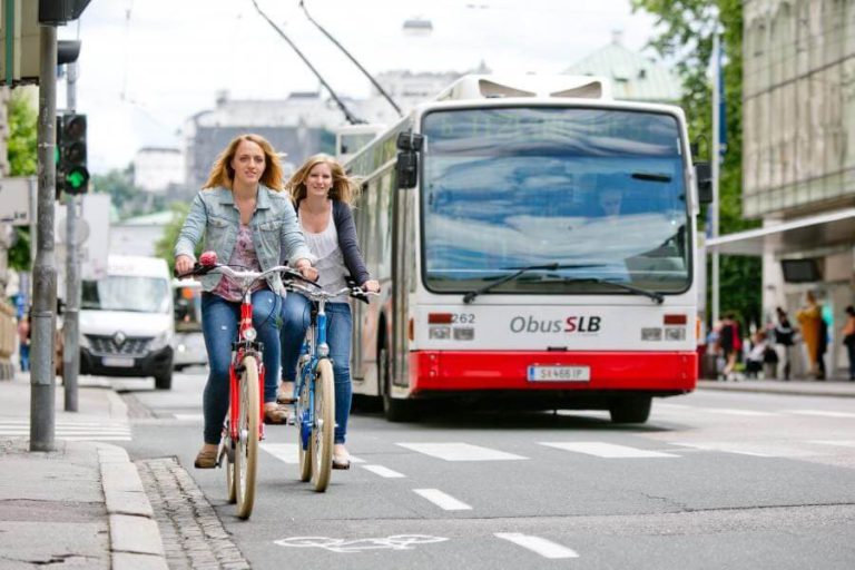 Zwei Frauen radeln auf einem Radweg in einer Stadtstraße, ein Bus fährt im Gegenverkehr.