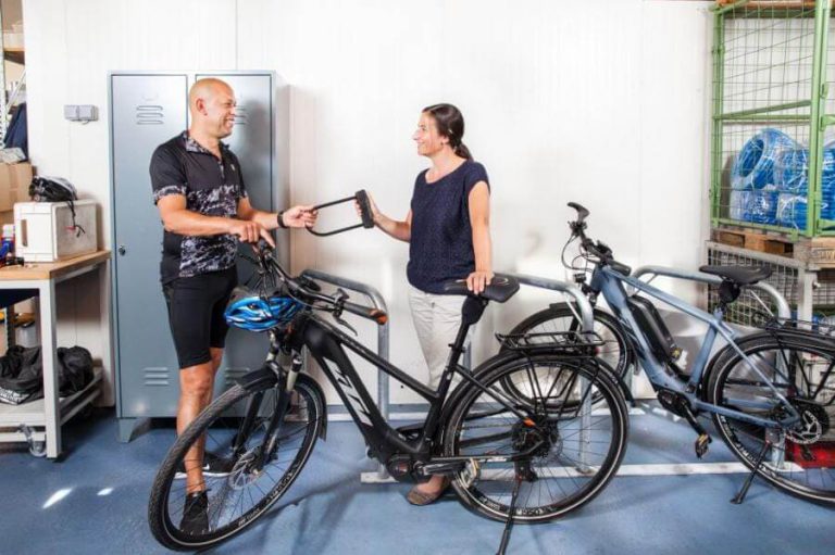 Zwei Personen übergeben sich ein E-Bike in einem Lagerraum mit weiteren Fahrrädern.
