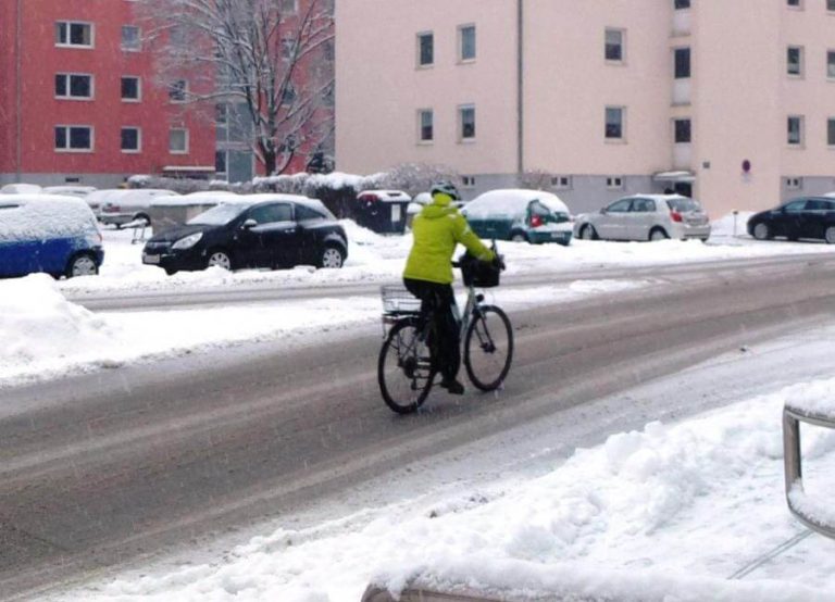 Radfahrer in gelber Jacke fährt auf schneebedeckter Straße in winterlicher Umgebung.