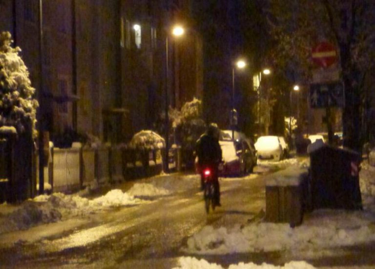 Radfahrer fährt nachts durch eine verschneite Straße, beleuchtet von Straßenlaternen.