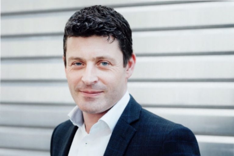 Mann in Anzug lächelt vor grauer Hintergrundwand, Thema: Klimaschutz & Mobilität.