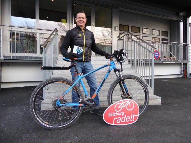 Mann mit Mountainbike und SalzburgRadelt-Schild vor einem Gebäude.