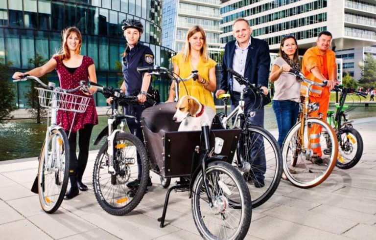 Sechs Personen mit Fahrrädern, darunter ein Lastenrad mit Hund, vor moderner Architektur.