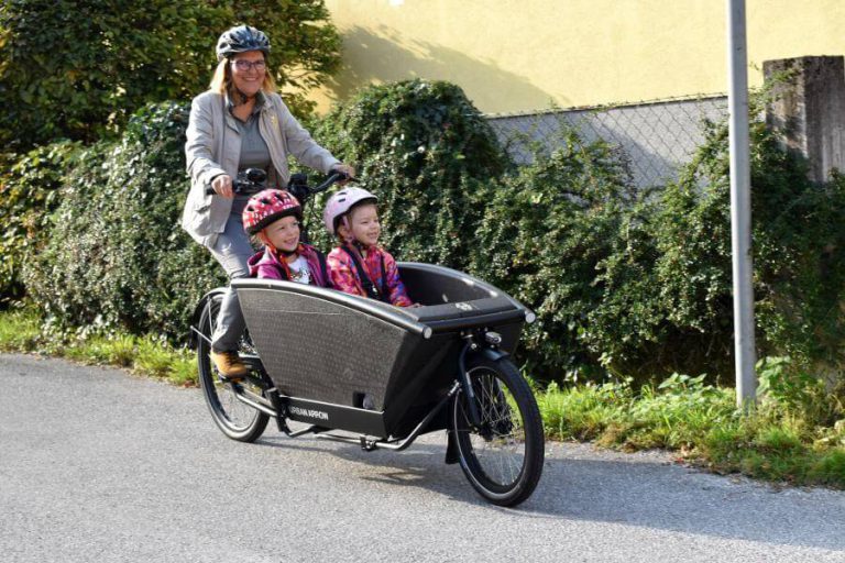Frau fährt mit zwei Kindern in einem Lastenrad entlang einer Straße in Salzburg.
