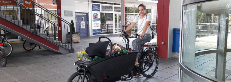 Frau fährt auf Longtail-Lastenrad mit Einkaufstüten und Blumen durch ein Einkaufszentrum.