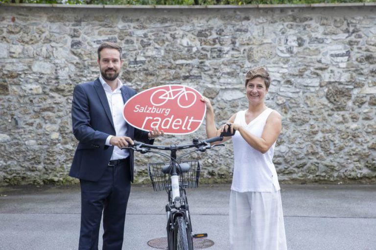 Julia Neudorfer und ein Mann halten ein „Salzburg radelt“-Schild vor einem Fahrrad.