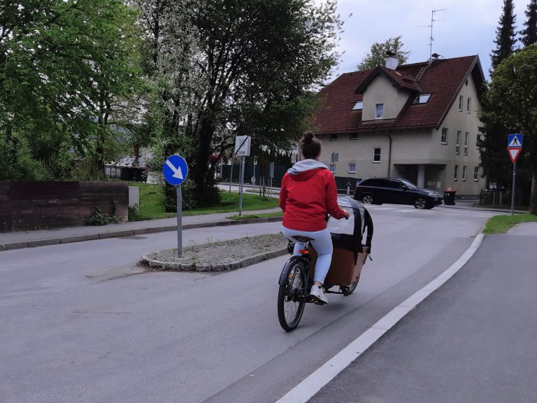 Frau mit Lastenrad auf Straße in Salzburg, umgeben von Bäumen und Häusern.