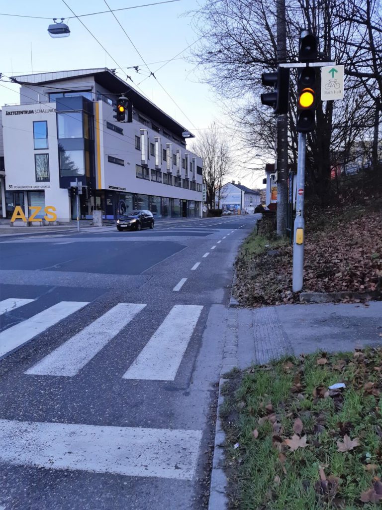 Ampel mit Fahrradweg-Piktogramm, Parkplatz und Ärztezentrum in Salzburg unmittelbar an der Straße.