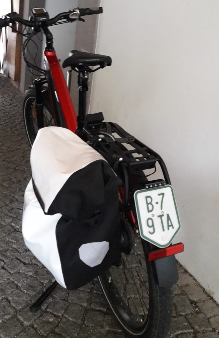 Rotes E-Bike mit Gepäckträger und Tasche in einem Gang auf Steinboden.