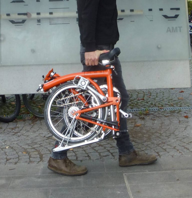 Mann trägt ein orangefarbenes, gefaltetes Fahrrad durch die Stadt auf einem gepflasterten Weg.