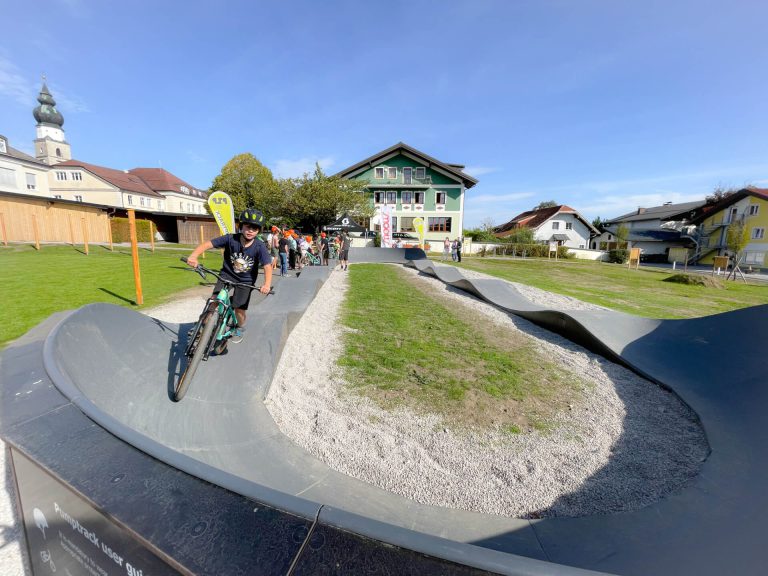 Kind fährt auf dem Pumptrack in Eugendorf, umgeben von Zuschauern und Wohngebäuden.