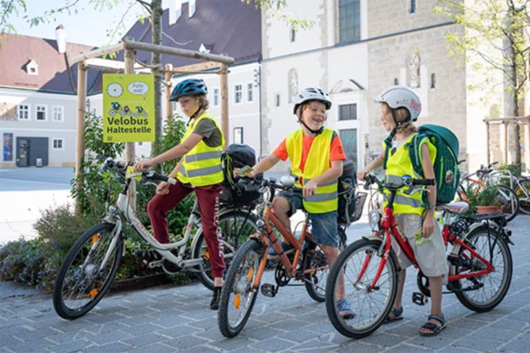 Drei Kinder mit Fahrradhelmen und Warnwesten stehen auf Fahrrädern an einer Haltestelle.