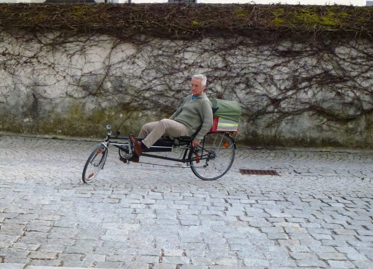 Senior auf einem Liegedreirad fährt entlang einer Steinpflasterstraße mit Weinstock-Wand.