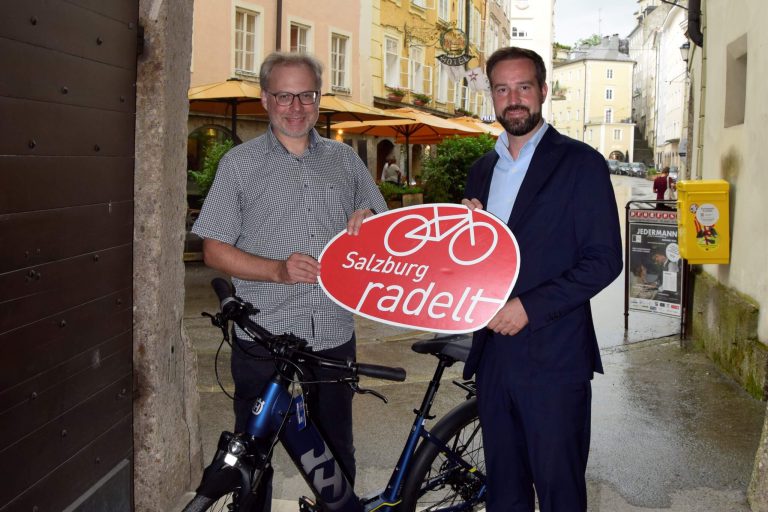 Zwei Männer mit Fahrrädern halten ein Schild „Salzburg radelt“ in einer Stadtstraße.