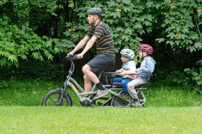 Vater fährt mit zwei Kindern auf einem Lastenrad durch eine grüne Wiese.