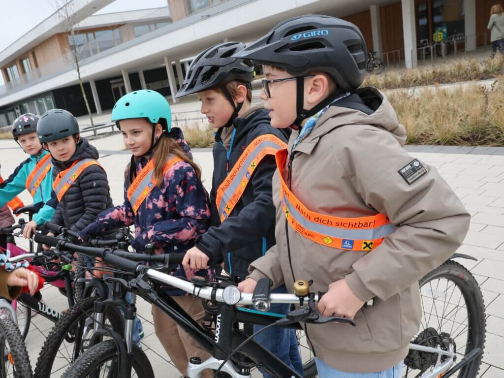 Kinder mit Helmen und leuchtenden Westen stehen neben ihren Fahrrädern, bereit zum Radfahren.