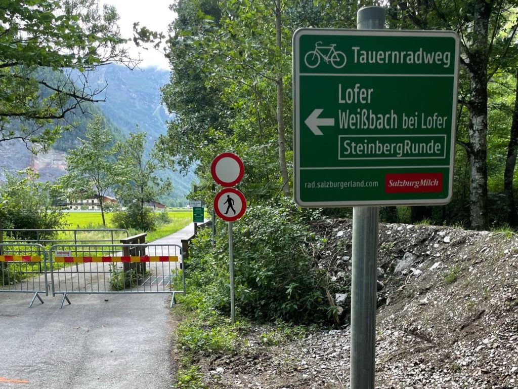 Wegweiser Tauernradweg Lofer, gesperrter Radweg, Berge und Wiese im Hintergrund