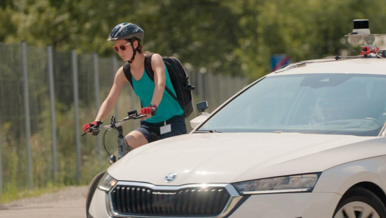 Radfahrerin fährt neben einem Auto mit Sensoren am Dach – Hinweis auf Totwinkelassistent und mehr Sicherheit im Verkehr
