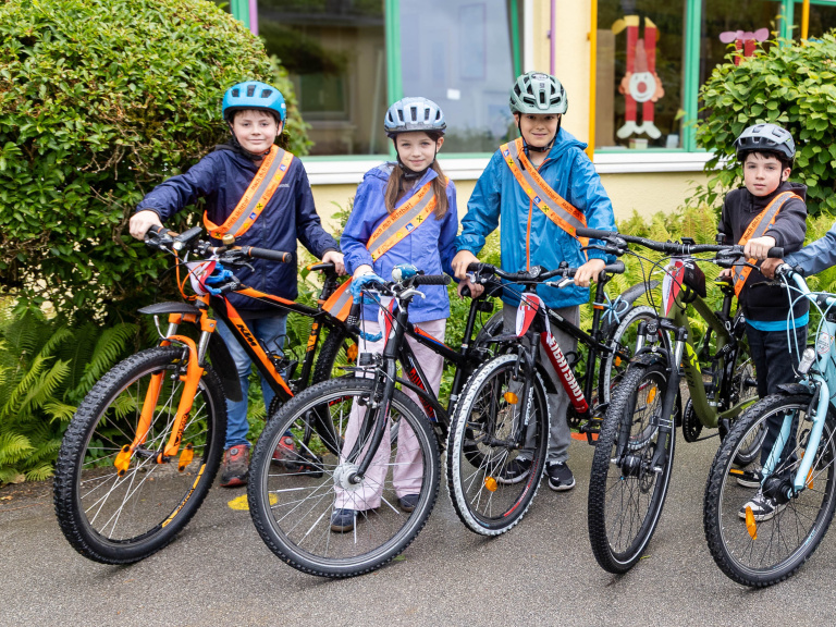 Vier Kinder mit Fahrradhelmen und reflektierenden Schärpen stehen mit Fahrrädern vor einem Schulgebäude