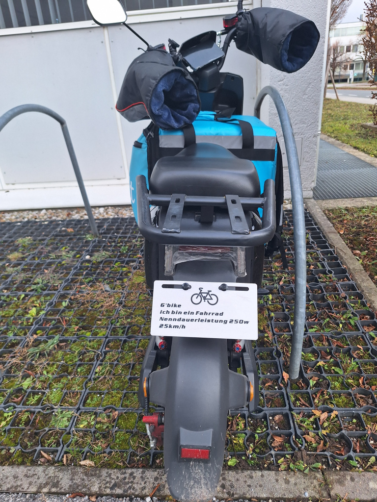 Geparkter Lieferroller mit blauer Transportbox und Hinweistafel neben einem begrünten Gitterstreifen