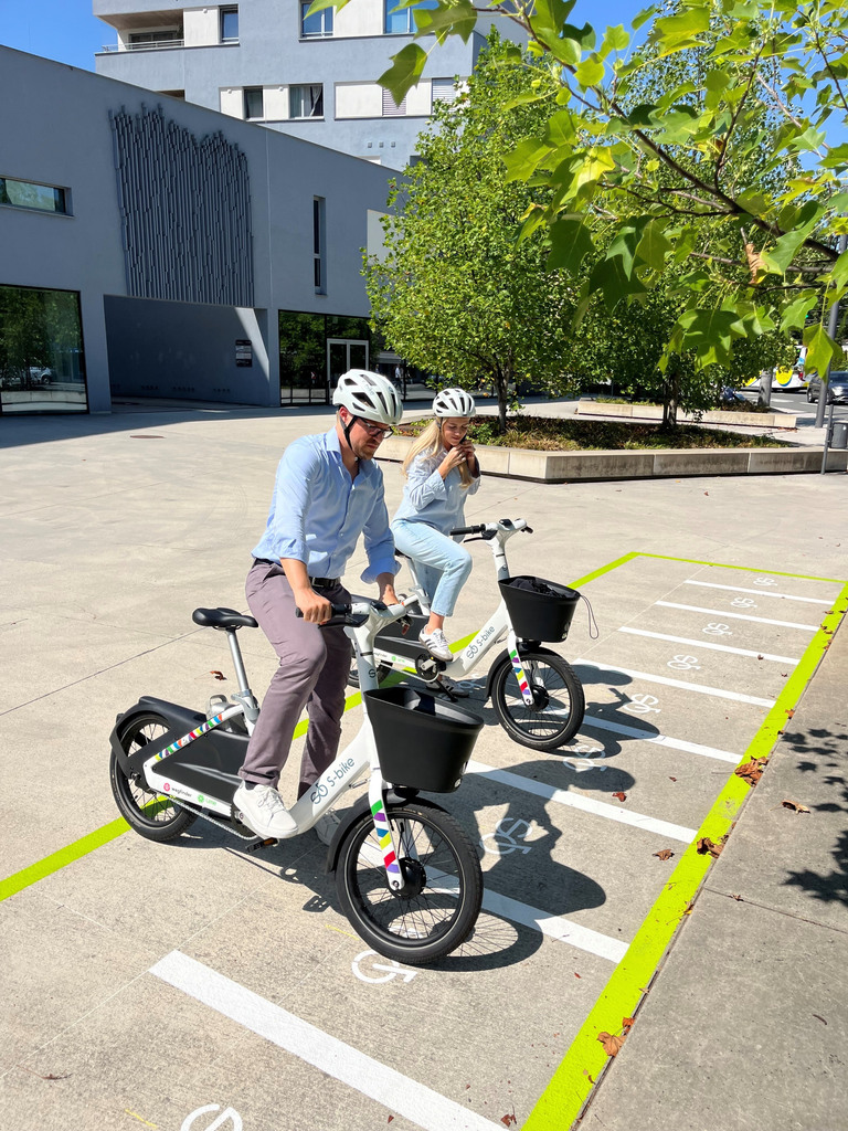 Zwei Helm tragende Personen auf weißen S-Bikes vor markierten Fahrradstellplätzen an einem modernen Gebäude